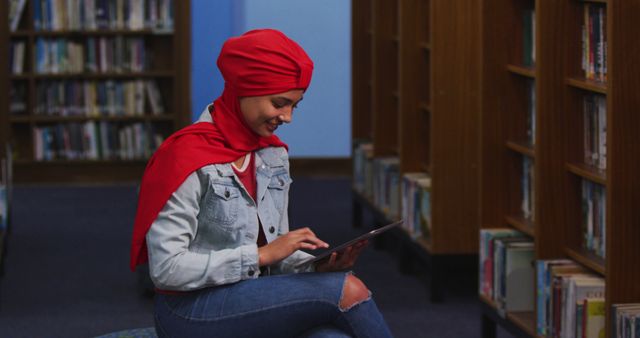 Young woman wearing red turban sitting on stool using tablet in a library, creating an image suitable for use in educational and technology-related content. The presence of bookshelves emphasizes the learning environment, making it ideal for articles or promotional material focusing on modern education, study habits, or digital learning resources. Perfect for illustrating the integration of technology in academic settings and diverse representations in learning spaces.