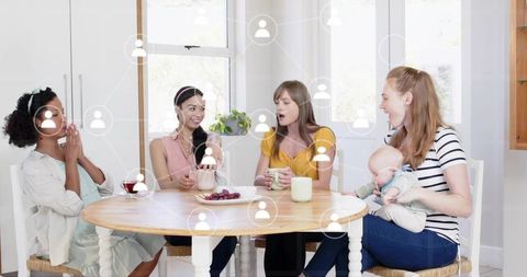 Moms chatting while holding mugs and baby at round wooden table in sunlit kitchen