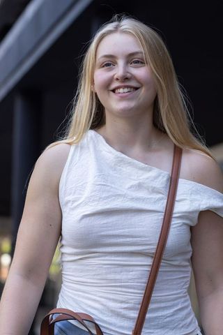 Young blonde woman smiling while walking urban sidewalk in white one-shoulder top