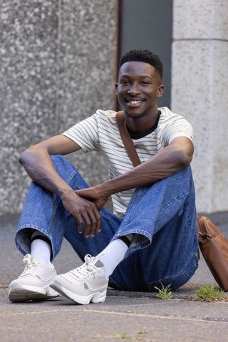 African man sitting on pavement smiling wearing striped t-shirt and backpack urban casual lifestyle