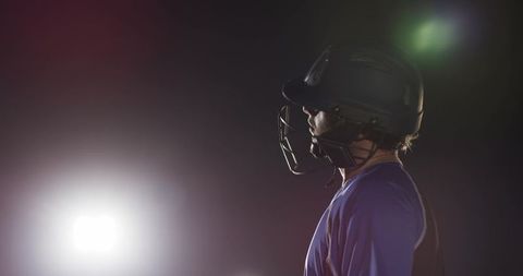 Standing baseball batter under stadium floodlights wearing helmet faceguard and jersey