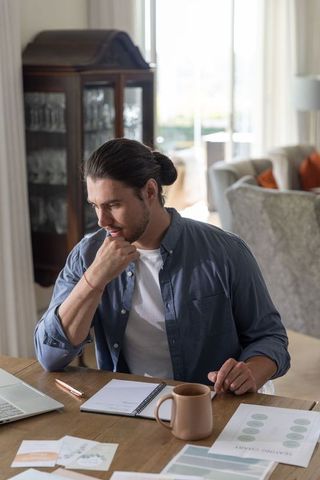 Man reviewing paperwork at home workspace, using laptop and notebook, coffee on desk, casual shirt