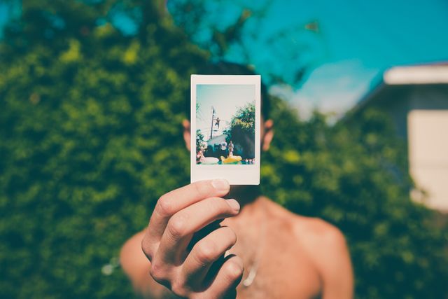 Man outdoors on a sunny day, holding Polaroid picture, face obscured by photograph. Perfect for use in promotions related to travel, summer activities, photography, and nostalgic moments. Highlights themes of memories, exploration, and outdoor leisure.