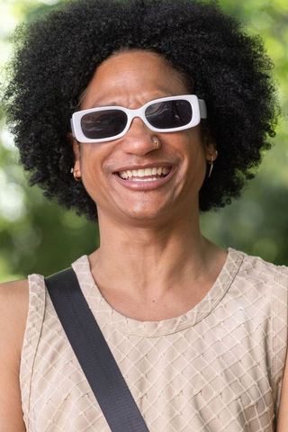 Smiling man wearing white rectangular sunglasses, gold nose hoop and earrings, beige top