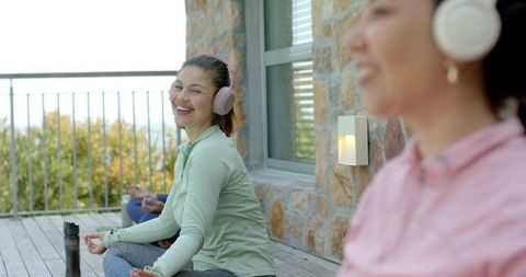 Multicultural women meditating on outdoor deck wearing headphones, practicing mindfulness