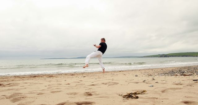 Man performing a martial arts kick on a sandy beach with a calming ocean background. Ideal for promoting fitness, martial arts training, mindfulness, summer activities, and wellness campaigns.