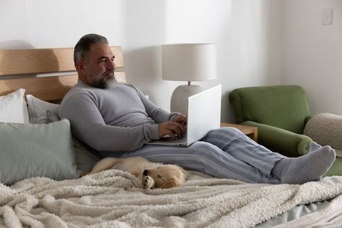 Mature man reclining on bed working on laptop with sleeping puppy in cozy bedroom