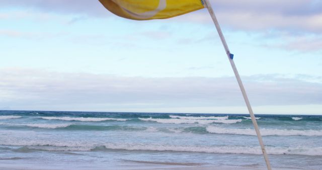 Yellow flag indicating surf conditions at beach with ocean waves and an overcast sky in the background. Perfect for coastal safety, lifeguarding, tranquil coastal scenes, and travel-related content.
