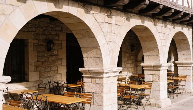 Sunlit stone arcade casting shadows over outdoor cafe with wooden tables, iron chairs, lamps