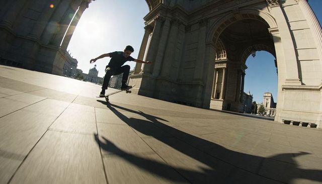 Skateboarder jumping under monumental stone arch casting long shadow with sun flare