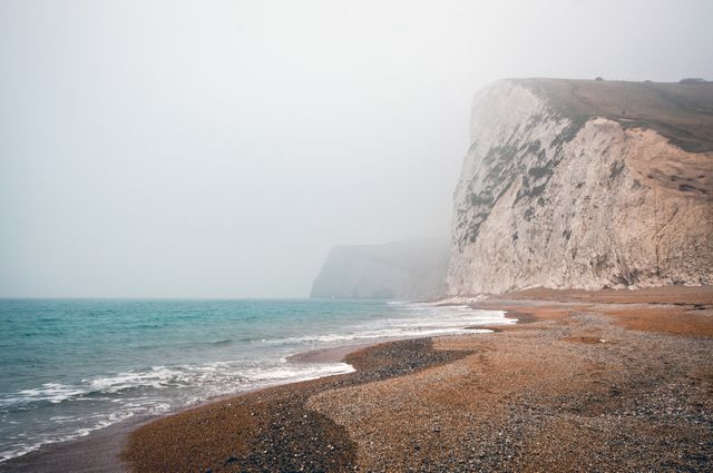 Picture capturing foggy coastal cliffs with pebble beach and calm sea, perfect for travel blogs, websites about nature, coastal landscapes, and serene escape destinations.