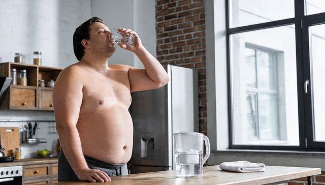 Shirtless man drinking water in industrial loft kitchen with filter pitcher for wellness