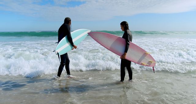 Two surfers in wetsuits holding surfboards are standing by the beach shore facing each other. They appear to be preparing for a surfing session. This visual can be used for content related to surfing, beach activities, outdoor sports, friendships, and ocean conservation.