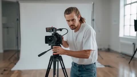 Videographer Adjusting Camera and Tripod in Studio Backdrop Preparing Professional Shoot
