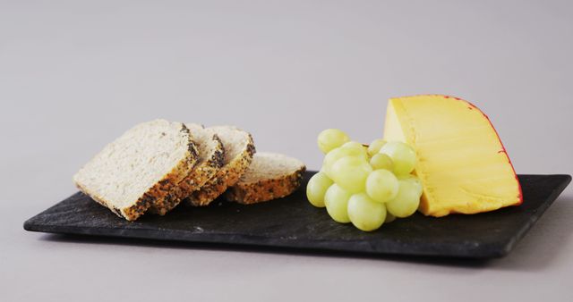 Slices of multi-grain bread, a wedge of cheese, and a bunch of green grapes are neatly arranged on a slate board, with copy space. The composition suggests a simple yet elegant snack or cheese platter setup.