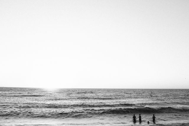 This black and white photo captures an ocean scene at sunset, with people standing in the water. The serene and calm atmosphere makes this perfect for use in travel, nature, and relaxation-themed projects. It is ideal for background images in websites, posters, or advertisements focusing on tranquility and natural beauty.