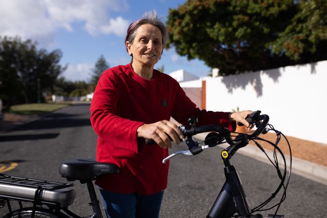 Senior woman with short grey hair wearing a red sweater is enjoying a sunny day while sitting on a bicycle in a residential street. Ideal for use in content related to active aging, healthy lifestyles, outdoor activities, retirement living, and promoting fitness among seniors.