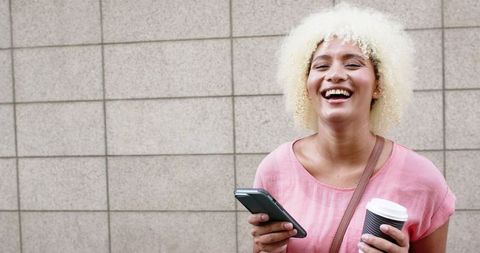 Joyful Young Woman Holding Smartphone and Coffee Against Urban Background