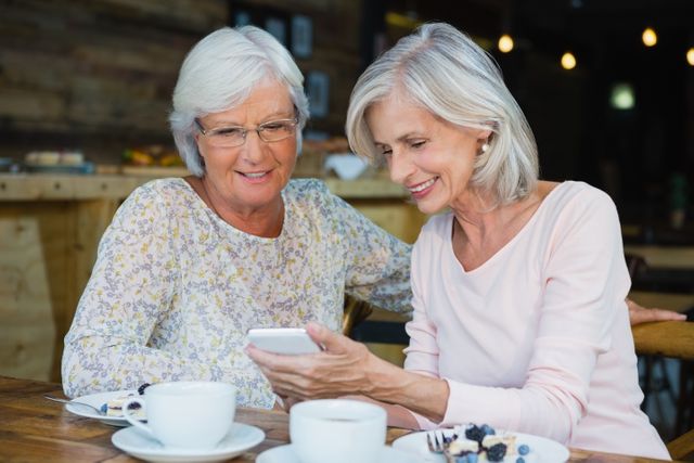 Two senior women are sitting at a cafe table, enjoying coffee and using a mobile phone together. They are smiling and appear to be sharing a moment of joy and connection. This image can be used for promoting senior lifestyle, technology use among elderly, social activities, and friendship themes.