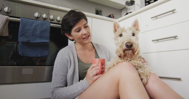 Woman sitting on kitchen floor with her dog, holding coffee mug. West Highland White Terrier on her lap. Cozy and casual moment in home setting. Ideal for themes related to pet care, home life, relaxation, companionship, and casual day-to-day moments.