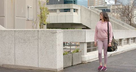 Mature woman walking with duffel on walkway in pink activewear and brutalist architecture