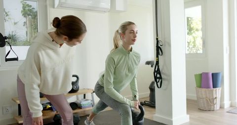 Female trainer guiding client performing kettlebell lunge in bright home gym with suspension straps