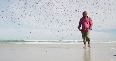 Senior man walking barefoot on beach, pink shirt, purple confetti drifting over calm ocean