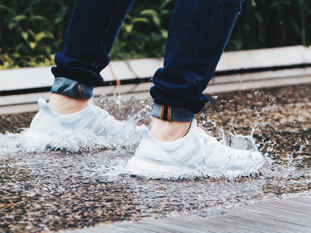 This image captures a close-up of white sneakers splashing through a shallow water puddle. Ideal for advertisements or articles about outdoor activities, comfortable casual footwear, urban lifestyle, or rainy day fashion tips. The action shot highlights movement and the water droplets, adding a dynamic element to the scene.