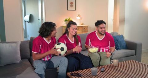 Friends cheering during soccer match on couch wearing matching red jerseys sharing popcorn