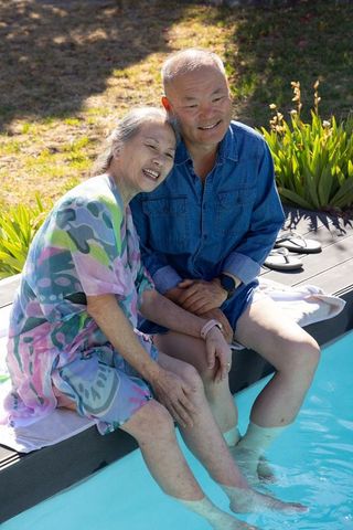 Senior asian couple relaxing by poolside dipping feet and smiling in sunlit backyard