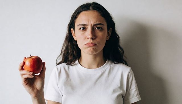 Somber young woman holding red apple in minimalist studio portrait off-white backdrop