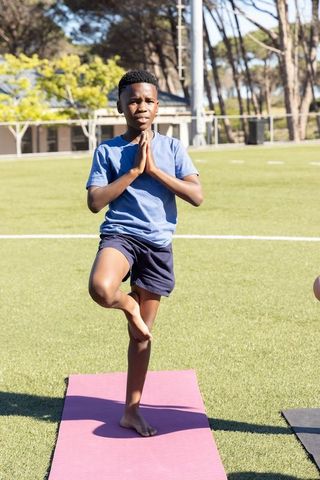 African american teen practicing tree pose on pink mat outdoors on school field for youth
