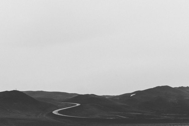 Winding road through a lonely mountain landscape captured in black and white. Can be used in creative projects that convey feelings of serenity, isolation, tranquility, or simplicity. Great for travel blogs, minimalist art, motivational posters, and editorial uses.