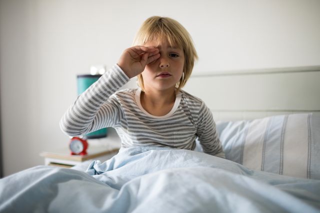 Young boy sitting in bed, rubbing his eyes, looking sleepy. Ideal for concepts related to morning routines, children's sleep habits, waking up, and home life. Can be used in articles, blogs, or advertisements about parenting, sleep health, and children's daily routines.