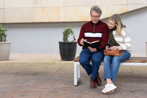 Senior couple sharing notes on bench, man writing notebook and woman checking smartphone