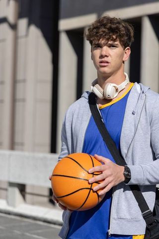 Teen athlete holding basketball on urban college campus wearing headphones and hoodie