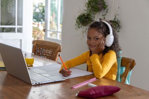 Young girl studying at home with laptop and headphones writing in notebook at dining table