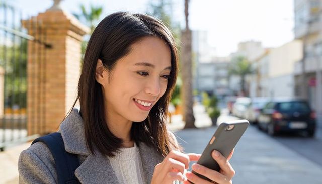 Smiling asian woman tapping smartphone while walking on sunny sidewalk wearing grey wool coat