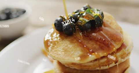 Pouring maple syrup over blueberry-topped pancake stack, golden breakfast drizzle close-up