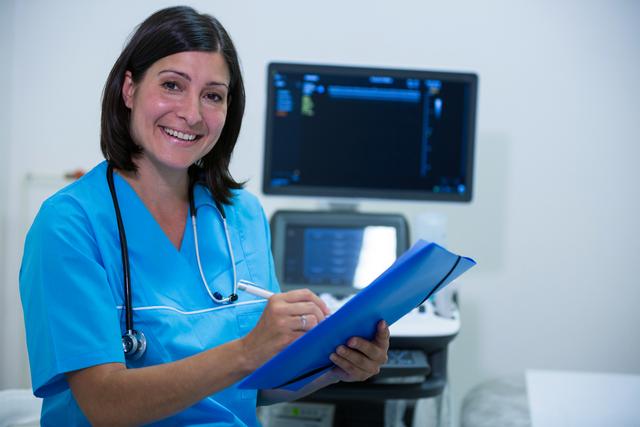 This image shows a smiling female doctor or nurse writing on a clipboard in a hospital room. She is wearing blue scrubs and has a stethoscope around her neck. Medical equipment is visible in the background. This image can be used for healthcare-related articles, medical websites, hospital brochures, and patient care promotions.