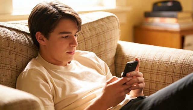 Teen texting on smartphone reclining on beige plaid couch in sunlit cozy living room