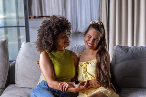 Smiling women couple holding hands on sofa, intimate home romance, smartwatch visible