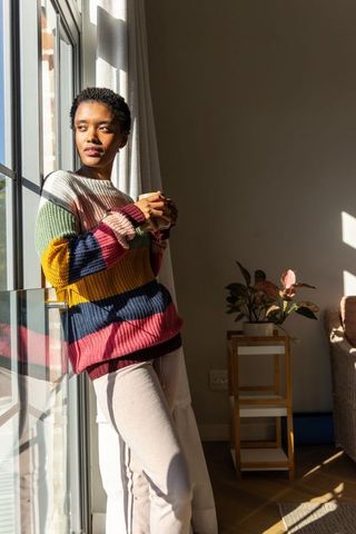 African american woman standing at window in striped sweater gazing into sunlight