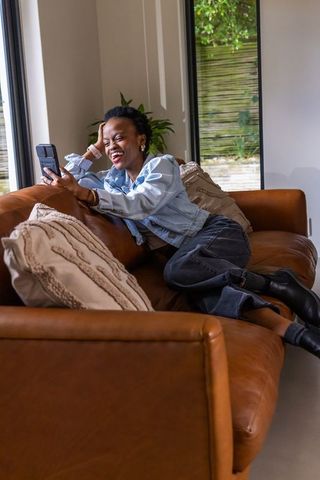 African american woman laughing on leather sofa using smartphone in modern living room