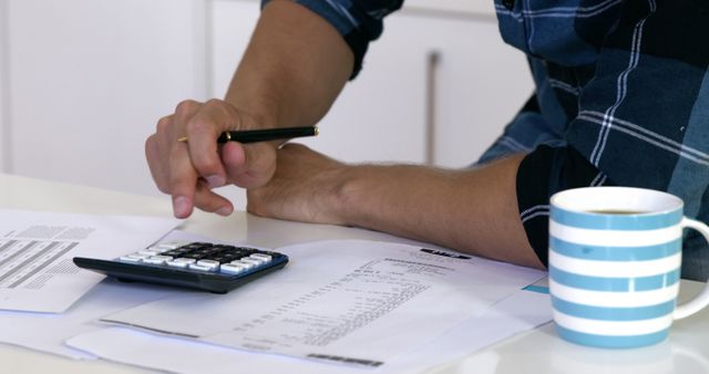 Person reviewing and organizing financial documents on a desk with a calculator and coffee. Ideal for use in articles about personal finance, budgeting tips, financial planning, remote work lifestyle, and home office productivity.