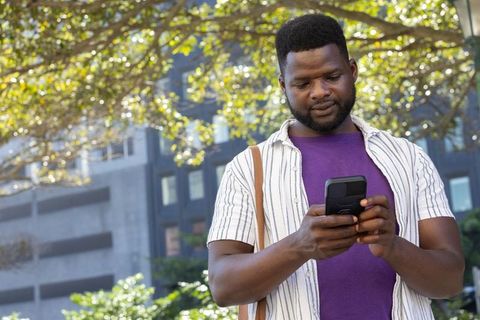 Urban commuter checking smartphone under sunlit tree near office building with shoulder bag