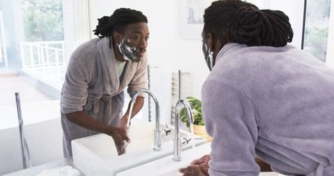 African American man washing face in plush robe at sunlit modern bathroom vanity