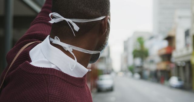 Man wearing protective mask looking down a city street, suggesting themes of urban living, health, safety, and anonymity. Suitable for use in articles or advertisements related to city life, personal protective equipment, public health, and safety.