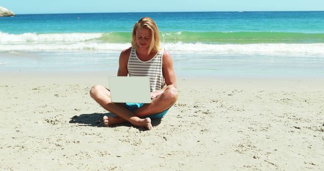 Man sits cross-legged on sandy beach by clear blue ocean, working on laptop. Ideal for themes related to remote work, digital nomad lifestyle, freelancing, technology in nature, beach vacation, summer work-life balance, and serene outdoor environments.
