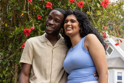 Sunlit diverse couple standing shoulder to shoulder smiling by red hibiscus shrub sharing joy